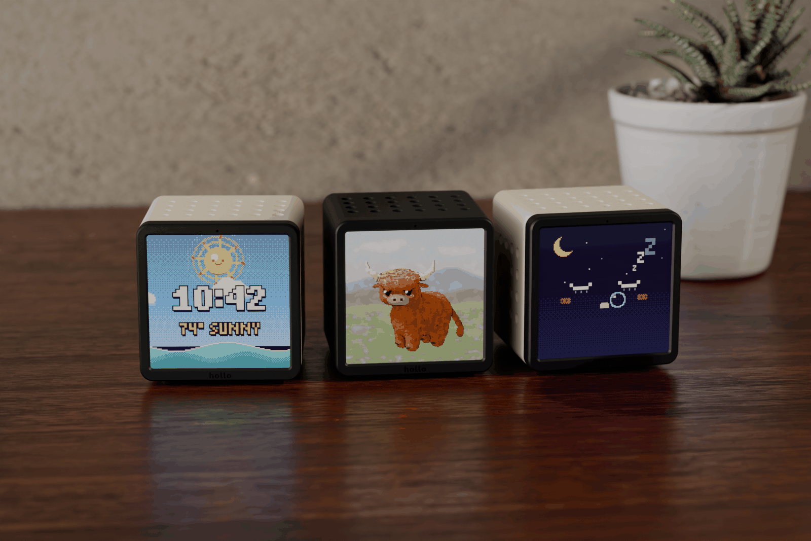 Rendered scene: three Hollo cubes side by side on a sunny desk, cream, black and cream, showing the clock with sunny weather, a pixel highland cow and a sleeping face